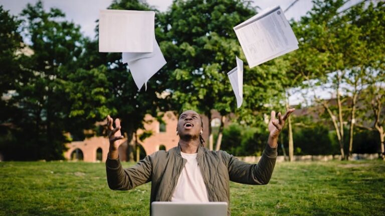 Happy young black male freelancer throwing papers while celebrating successful project during remote work in green park