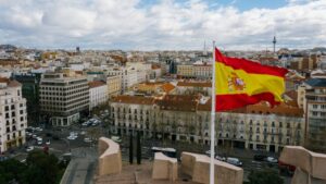 Drone view of Spanish city with aged buildings and national flag under cloudy blue sky