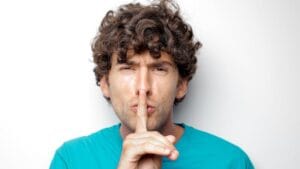 Adult man with curly hair in blue shirt making a silence gesture on white background.