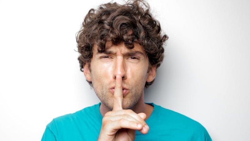 Adult man with curly hair in blue shirt making a silence gesture on white background.